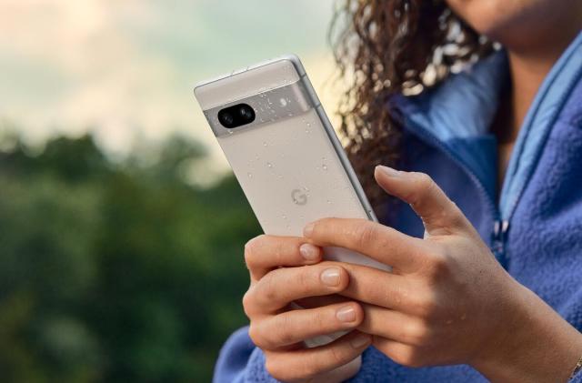 A person holds the snow-colored version of the Google Pixel 7a smartphone. There are water drops on the phone to illustrate its water resistance. 