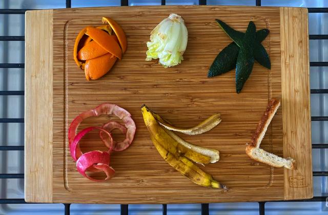 Food scraps, including lettuce, sandwich crust and peels from an orange, apple and banana, are arranged on a bamboo cutting board. 