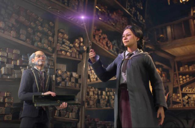 In this still from the video game 'Hogwarts Legacy': Surrounded by towering shelves holding long, thin boxes, a young witch in a Hogwarts school robe holds a wand aloft, its tip illuminated, as the shopkeeper looks on.