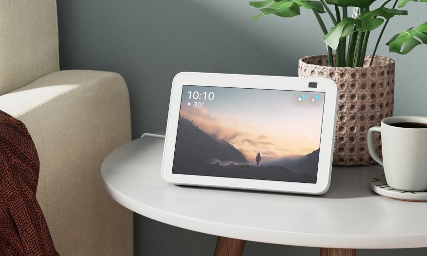 A white Amazon Echo Show 8 displays the time and temperature while resting on a side table in front of a potted plant and cup of coffee, next to a white couch.