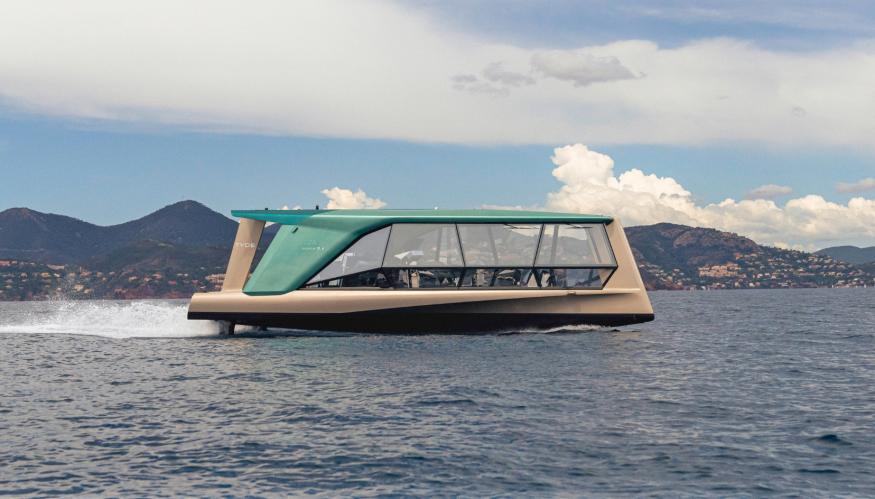 A side view of the glass-walled TYDE/BMW collaborative boat, called the Icon, as it moves across the water with hills and clouds in the distance.