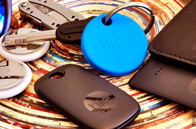 An assortment of Bluetooth trackers on a colorful background with a circular pattern. Trackers include a round blue Chipolo key chain tag, two white and silver AirTags, a black key-fob Tile tracker and a black wallet with a card-shaped tracker poking out. 
