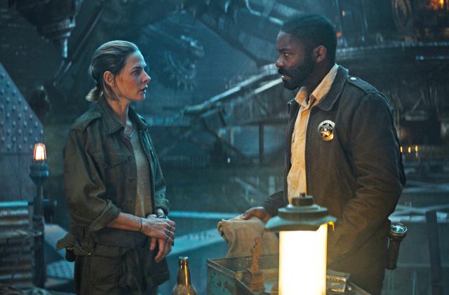Rebecca Ferguson and David Oyelowo stand in a darkened industrial space with large steel girders and machines in this promo image for the Apple TV+ series 'Silo.'