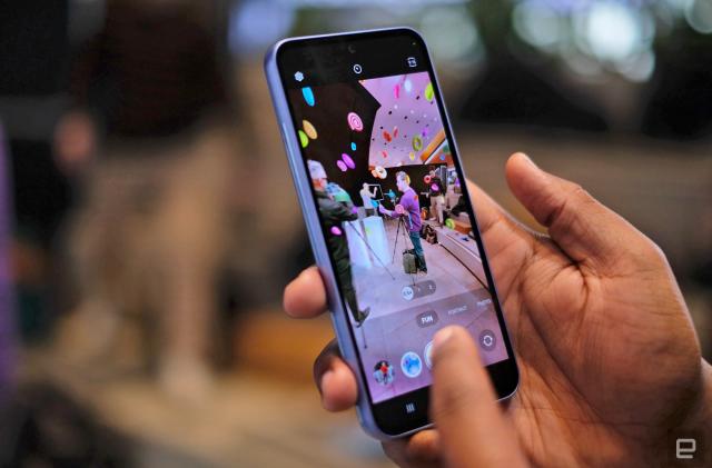 A cropped image shows two hands holding up a Samsung Galaxy A53 smartphone with a colorful event being shown on the phone's screen as the camera is activated.