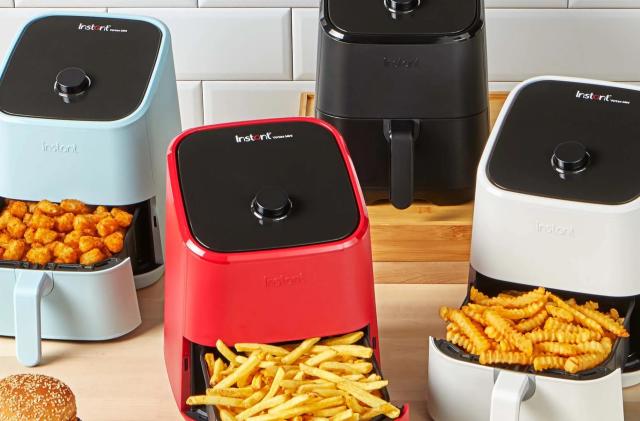A collection of compact air fryers in various colors sit on a wooden countertop against a white tiled wall, with their bottom compartments open, revealed cooked snacks like french fries and tater tots.