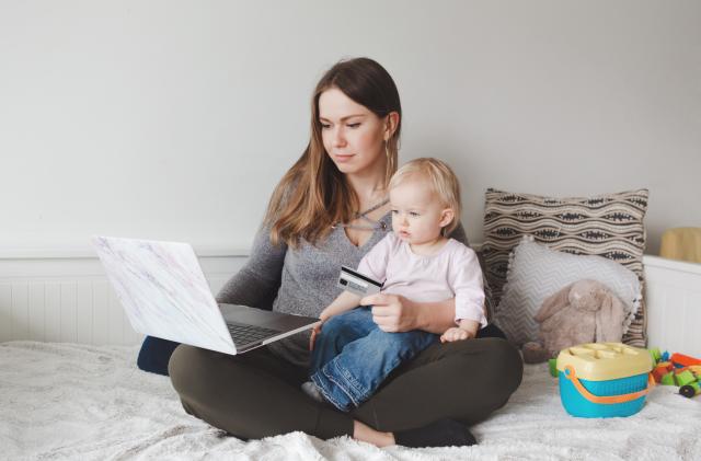 Young Caucasian mother with baby shopping online from home. Stay at home single mom with kid toddler daughter buying ordering products food on Internet and paying with credit card.