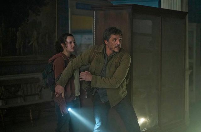 A promo image with Pedro Pascal and Bella Ramsey in HBO's The Last of Us. The two huddle in a dark corner of the room with a single flashlight, worried about some danger in the vincinity.