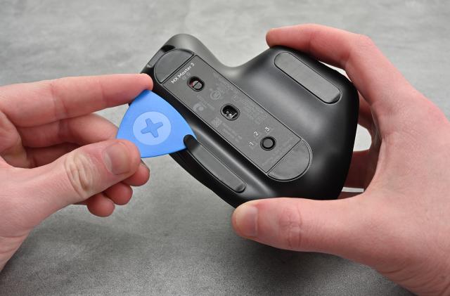 Photo of a person holding the Logitech MX Master 3 mouse with a blue spudger in the other hand pulling open a tab to open the device.