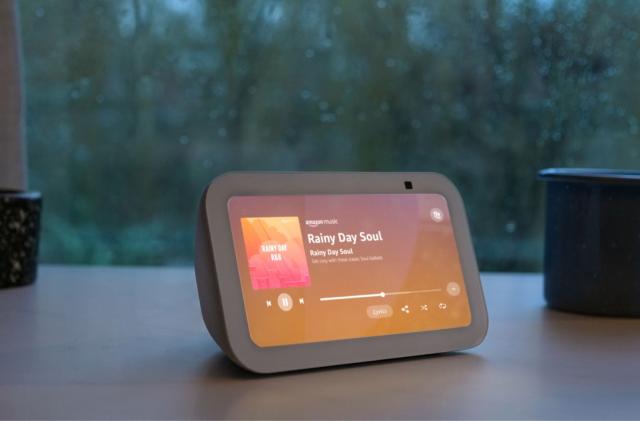 Amazon marketing photo of the Echo Show 5 (third-gen). The smart speaker has a display showing "Rainy Day Soul" on Amazon Music. It sits on a brown desk by a window with rain outside.