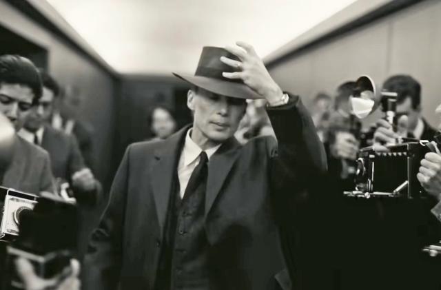 Cilian Murphy in 'Oppenheimer' walks past journalists with cameras in a hallway.