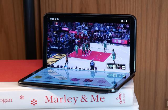 The Google Pixel Fold in "tabletop view" half opened atop a stack of books. A basketball game is displayed on the upper screen. Apps and other icons are visble on the bottom screen. 