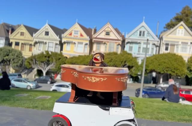 The muppetized Vanessa Carlton robot in a small wheeled vehicle with a piano on Steiner St in San Francisco.
