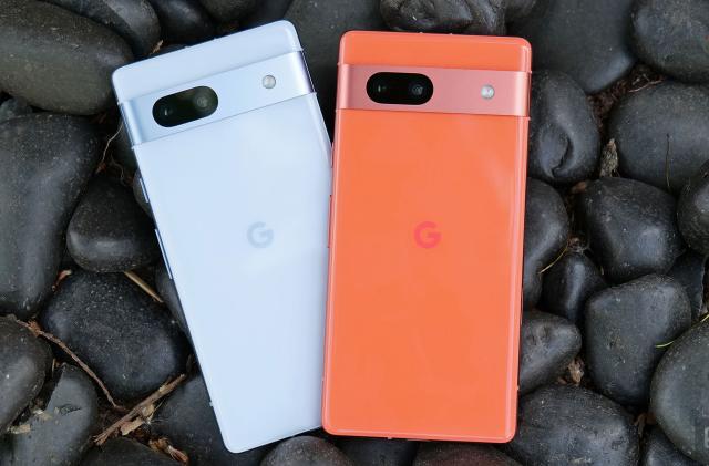 The sea and coral colored Pixel 7a phones sit on a bed of black rocks. 