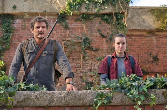 Pedro Pascal and Bella Ramsey stand behind a short brick wall and look at the camera in this promotional image from the TV series 'The Last of Us'.