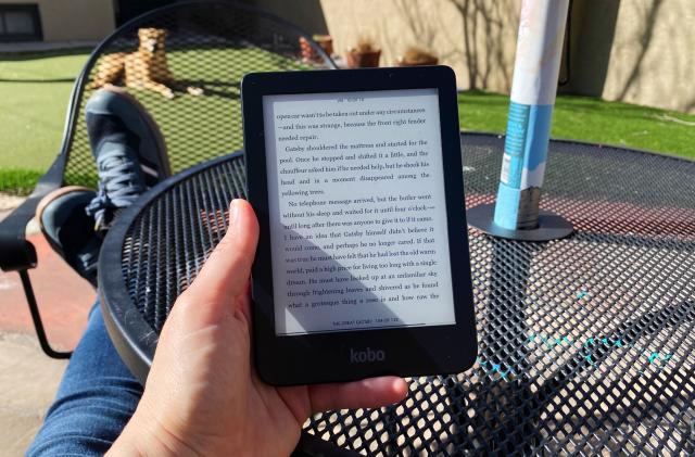 A person reads a Kobo ereader outside with their feet up. There's a black metal table in the foreground and patch of grass with a dog on it in the background. The text is from the public domain novel The Great Gatsby. 