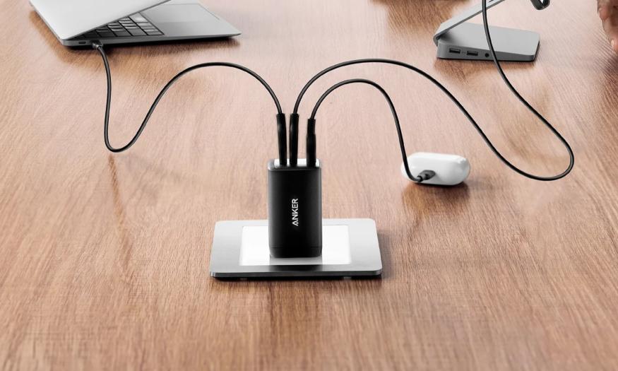 A small black wall charger, the Anker 735, shown plugged into an outlet on a brown wooden desk, with cables attached charging a laptop, smartphone and pair of wireless earbuds.