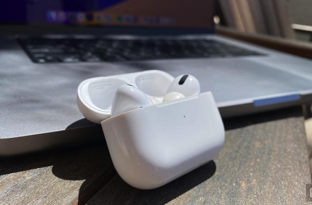 A pair of AirPods in the case sit on a wooden table next to an open MacBook. 